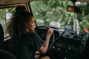 A person with curly hair sits in the driver's seat of a vehicle, steering while surrounded by greenery and personal items on the dashboard.