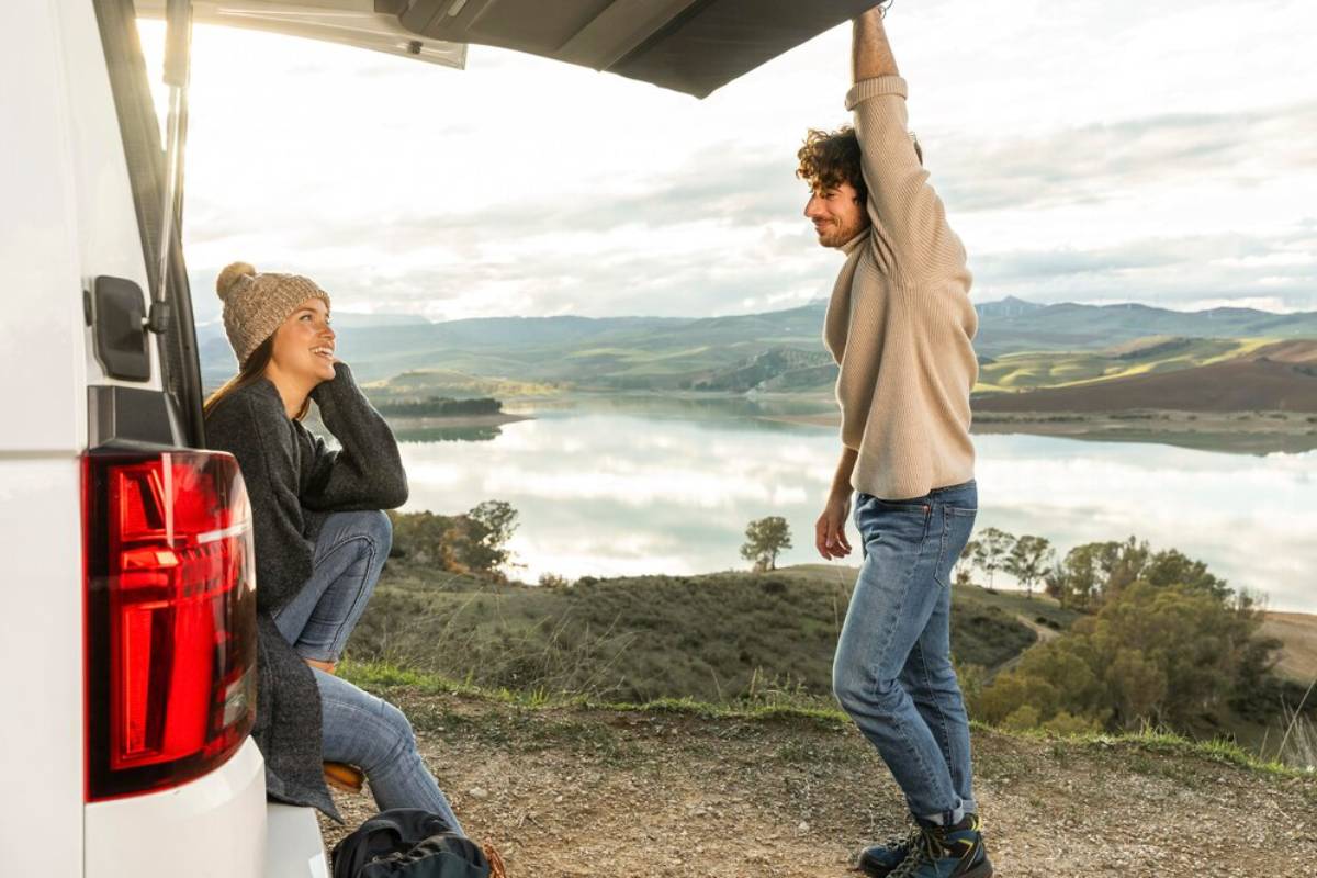 A couple enjoys the serene outdoors near a van, with a scenic lake and hills in the backdrop, capturing a moment of connection.