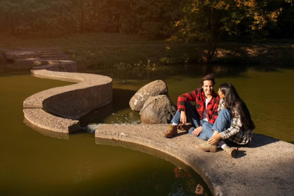 A couple sits together on a winding concrete pathway over still green water, surrounded by lush trees in warm sunlight.