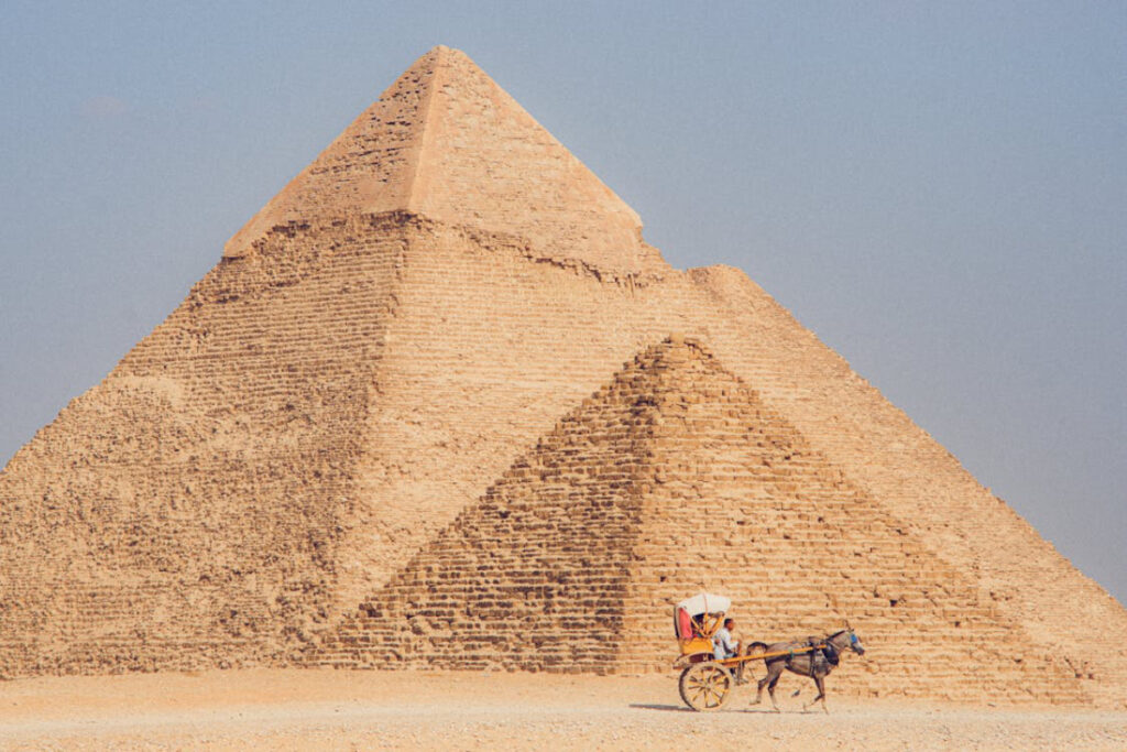 A horse-drawn cart with a person travels past a grand, ancient pyramid against a clear blue sky in Egypt.