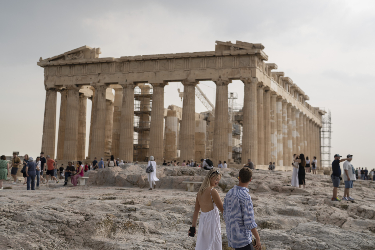Visitors explore the Acropolis, with the partially restored Parthenon towering in the background against a cloudy sky.