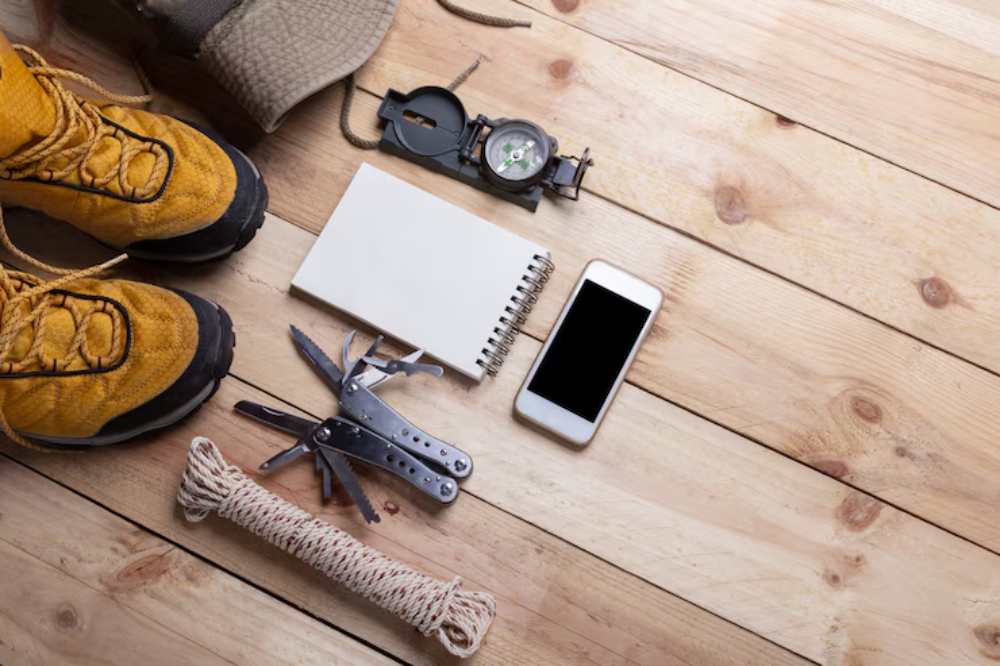 Flat lay of hiking gear: yellow boots, compass, multi-tool, notebook, smartphone, and rope on a wooden floor background.