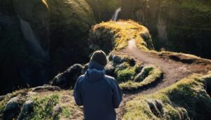 A hiker in a gray jacket and green hat gazes at a lush, winding path overlooking a scenic canyon and waterfall.