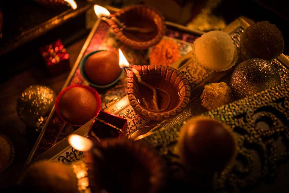 Diwali celebration scene with lit clay lamps and assorted sweets on ornate trays. The warm glow creates a festive and cozy atmosphere.