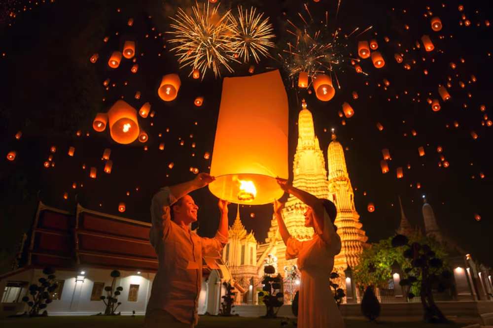 Couple releases a glowing lantern into a night sky filled with floating lanterns and vivid fireworks. Ornate temple towers are illuminated in the background.