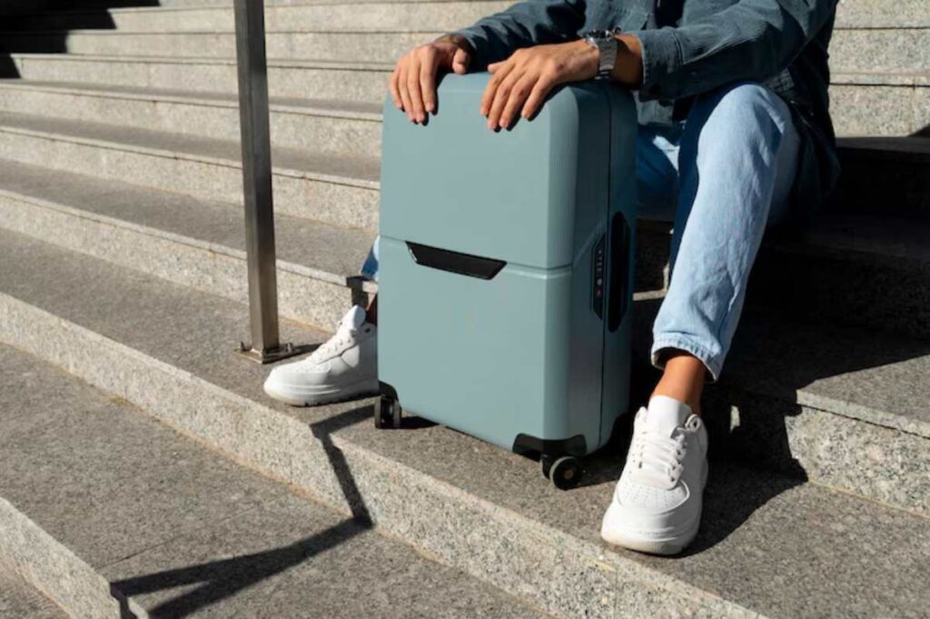 A person in a denim jacket sits on granite steps beside a light blue smart suitcase and wears white sneakers.