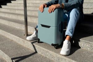 A person in a denim jacket sits on granite steps beside a light blue smart suitcase and wears white sneakers.