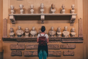 A person views a collection of classical marble busts displayed on a wall, surrounded by ornate stone carvings in an Open-Air Museum.