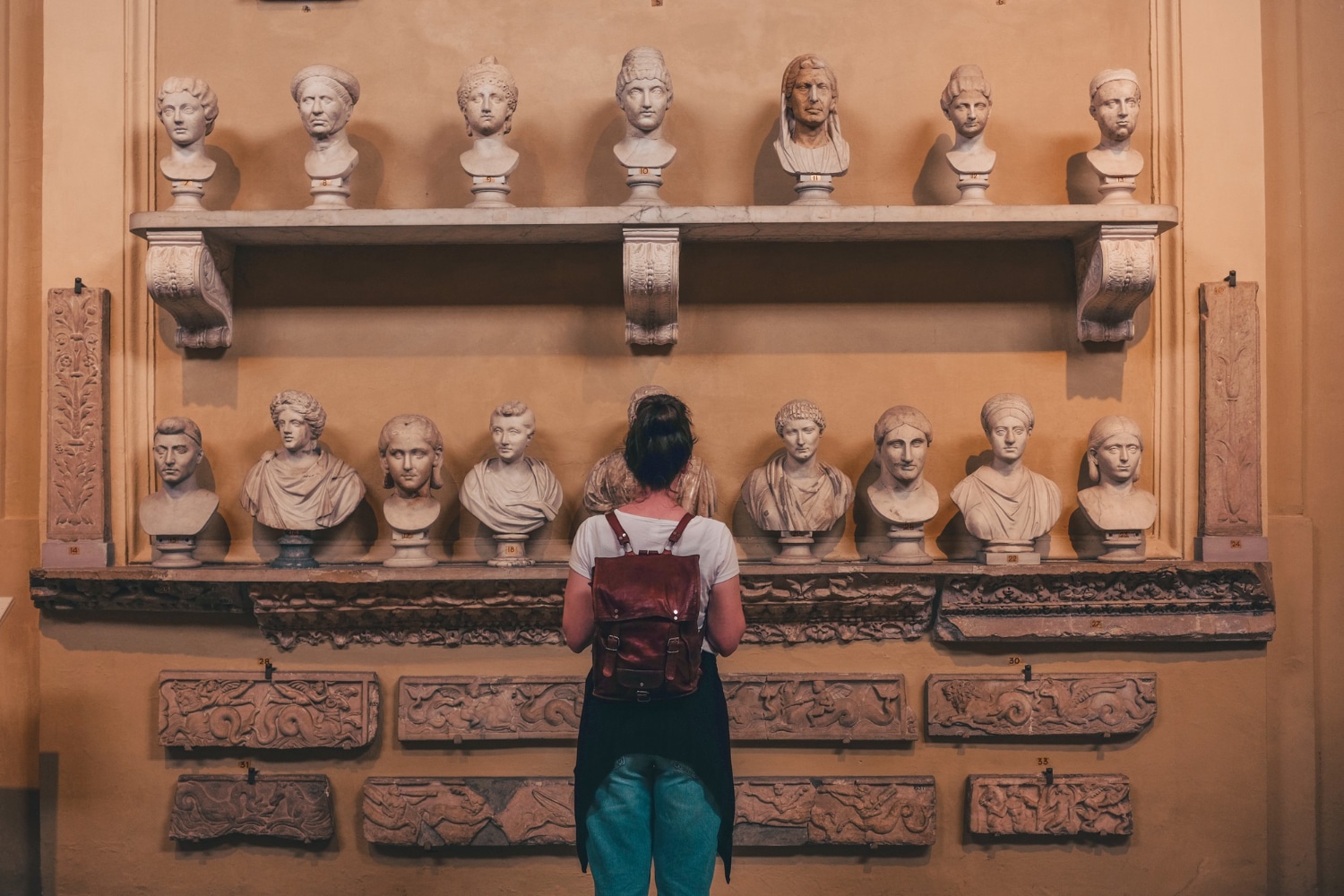 A person views a collection of classical marble busts displayed on a wall, surrounded by ornate stone carvings in an Open-Air Museum.