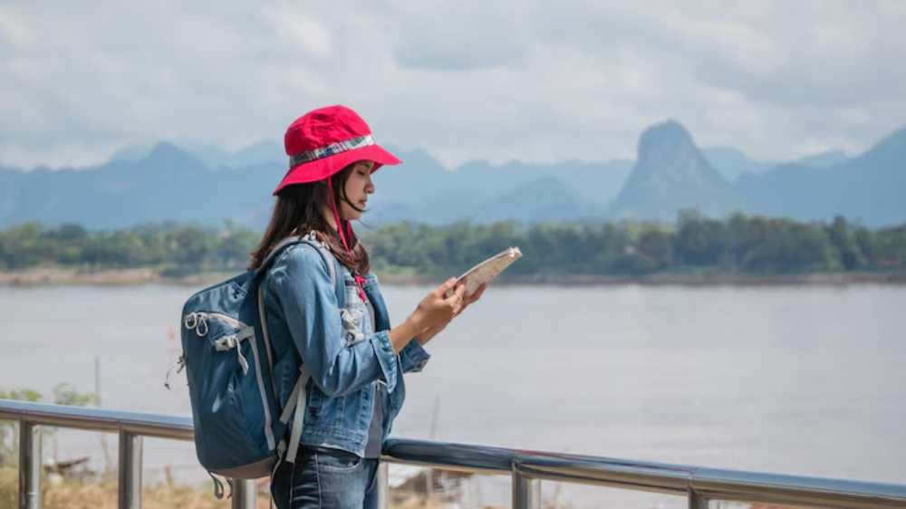 A traveler in a red hat and denim jacket stands by a river, reading a guidebook with mountains in the background.
