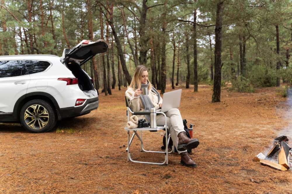 A person sits in a foldable chair and there is a car nearby, working on a laptop with a drink in hand.