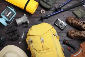 A yellow backpack surrounded by camping gear: a lantern, binoculars, camera, compass, hiking boots, and a yoga mat on a wooden surface.