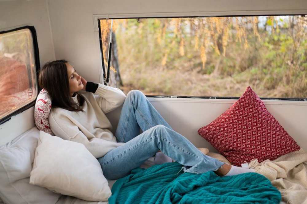 A person relaxes inside a cozy van, surrounded by pillows and a blanket. 