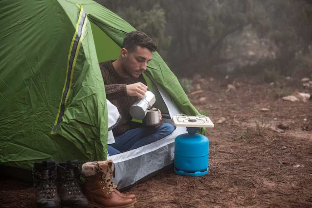 A person sits at the entrance of a tent, preparing coffee on a portable stove next to a blue gas canister.