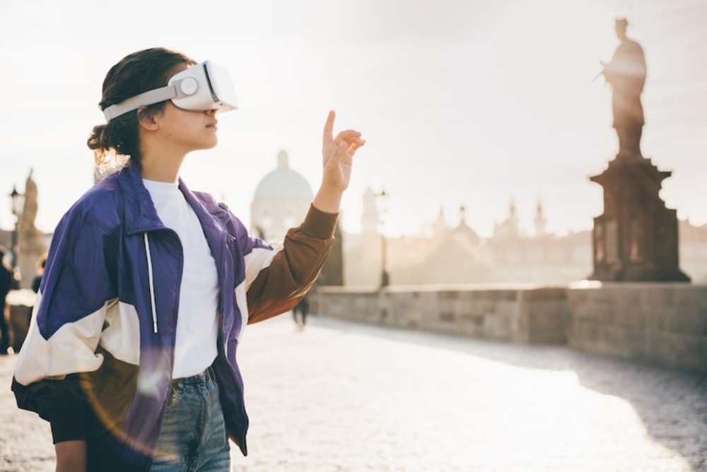 A person wearing a VR headset  standing in a sunlit urban environment with historic statues in the background. 